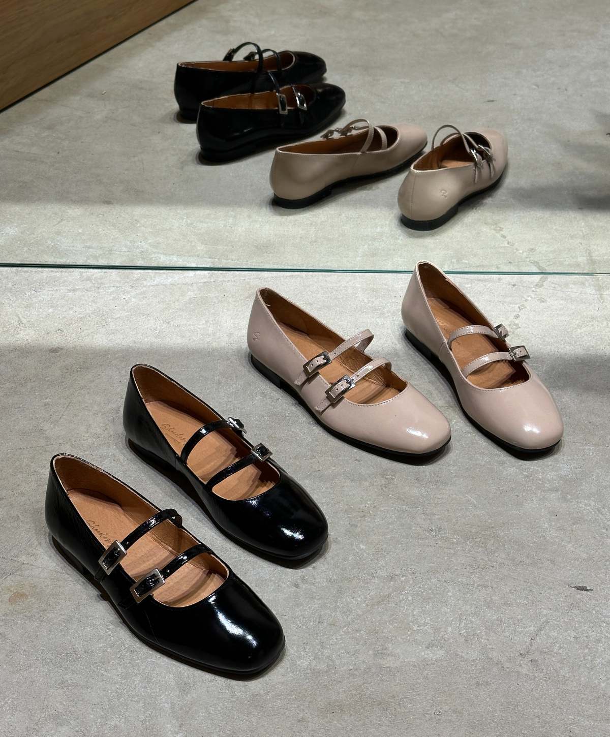 Sort og beige Mary Jane-sko med spænder, placeret på en grå betonoverflade foran et spejl, der reflekterer skoene.