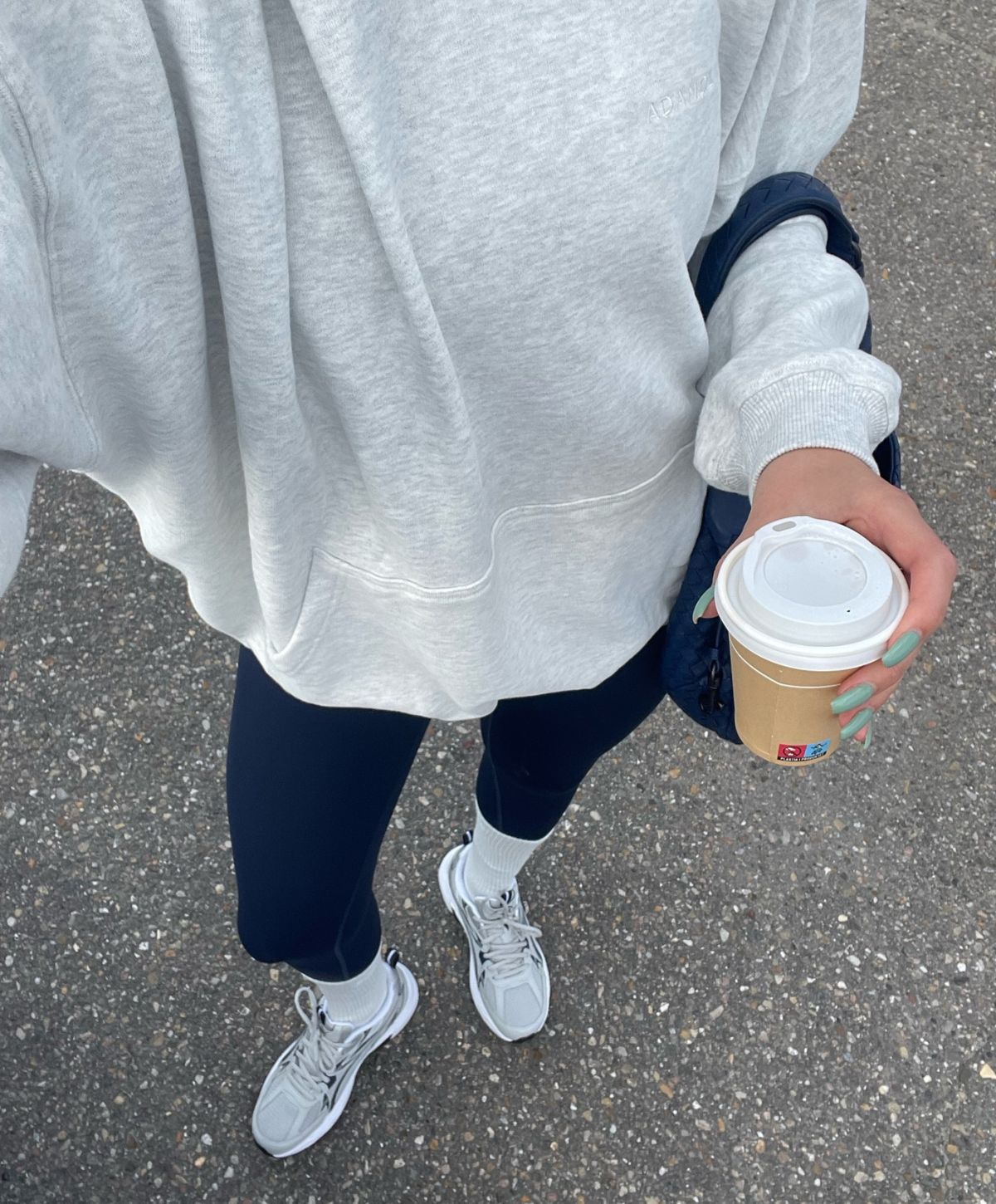 En person iført en grå sweatshirt, mørke leggings og hvide sneakers holder en papirkop med drikke.