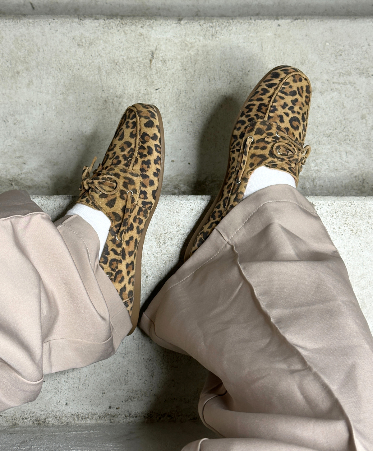 Brune leopardmønstrede loafers på fødderne af en person iført lyse beige bukser, der sidder på en betontrappe.