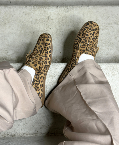 Brune leopardmønstrede loafers på fødderne af en person iført lyse beige bukser, der sidder på en betontrappe.