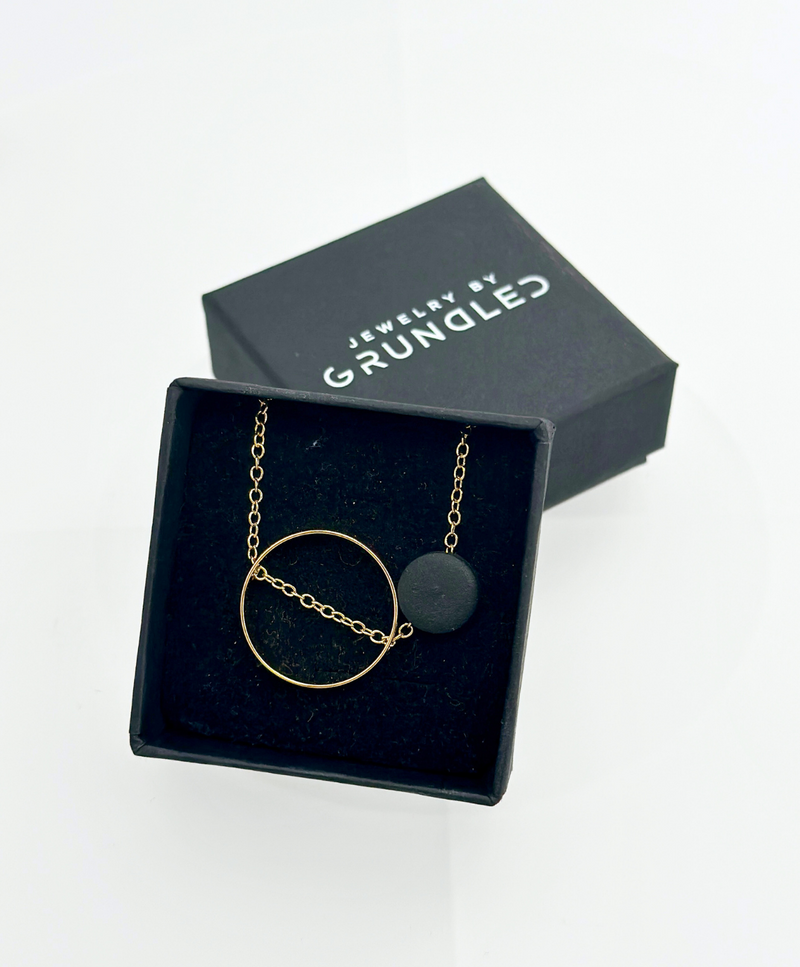 Sort smykkeæske med teksten "Jewelry by Grundled" indeholder en halskæde med en gylden cirkel og en sort rund vedhæng.